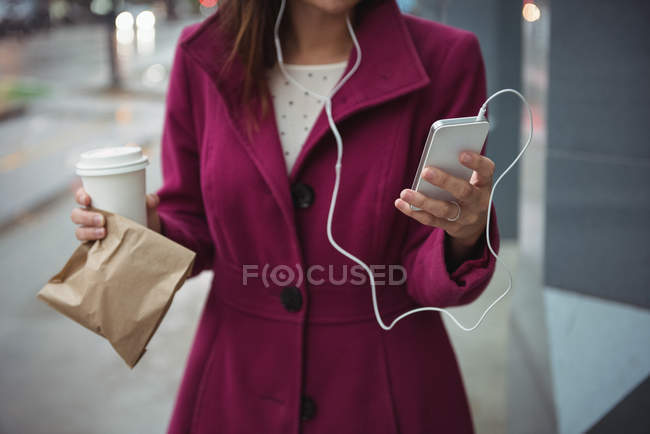 Businesswoman holding disposable coffee cup and parcel while listening to music near office building — Stock Photo