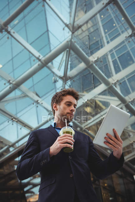 Geschäftsmann benutzt digitales Tablet, während er in der Nähe von Bürogebäuden Saft trinkt — Stockfoto