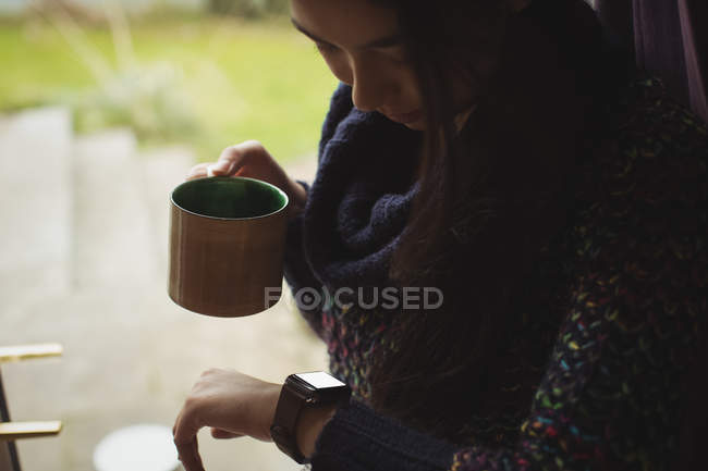 Mulher olhando para relógio inteligente enquanto toma café em casa — Fotografia de Stock