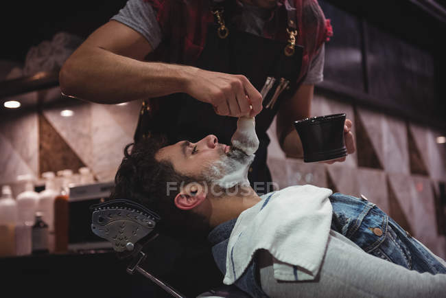 Средняя часть парикмахера наносит крем на бороду клиента в парикмахерской — стоковое фото