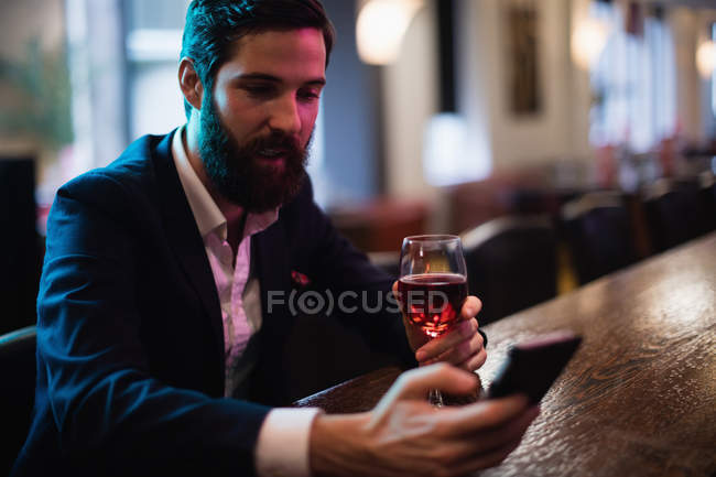Homme d'affaires utilisant un téléphone portable avec un verre de vin rouge à la main au bar — Photo de stock