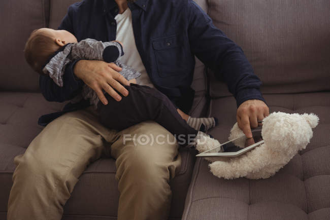 Vater hält sein Baby während er digitales Tablet auf dem Sofa benutzt — Stockfoto