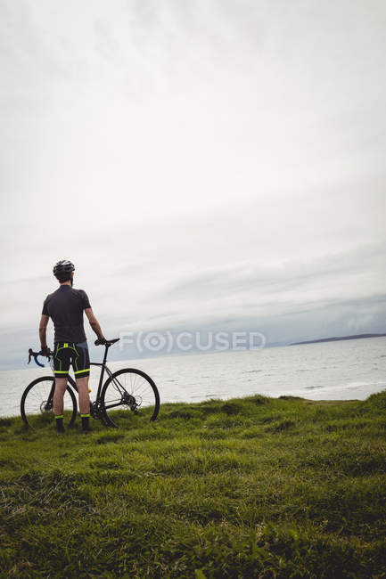 Atleta de pé com sua bicicleta na grama perto do mar — Fotografia de Stock