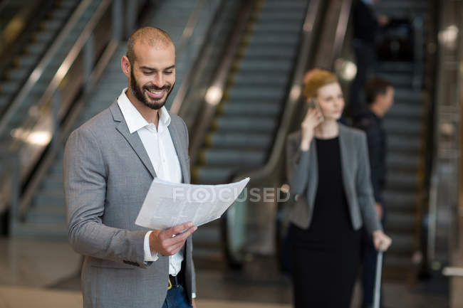 Hombre de negocios sonriente parado en la sala de espera leyendo el periódico en la terminal del aeropuerto - foto de stock