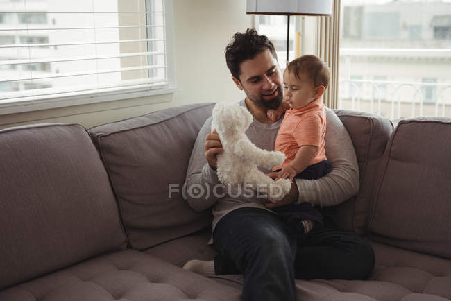 Отец и ребенок играют с плюшевым мишкой на диване в гостиной дома — стоковое фото