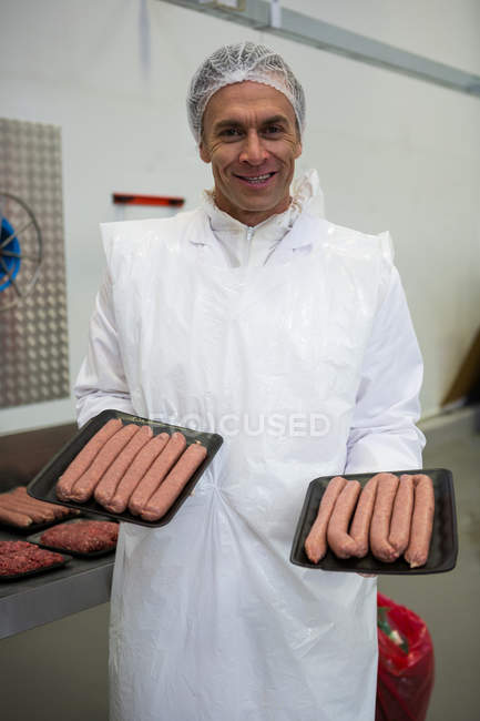 Retrato del carnicero sosteniendo bandejas de embutidos de carne en la fábrica de carne - foto de stock
