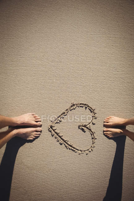 Ноги пари, що стоять біля серця намальовані на пляжному піску — стокове фото