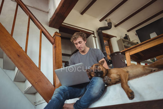 Homem sentado no degrau e usando laptop em casa, cão deitado ao lado dele — Fotografia de Stock