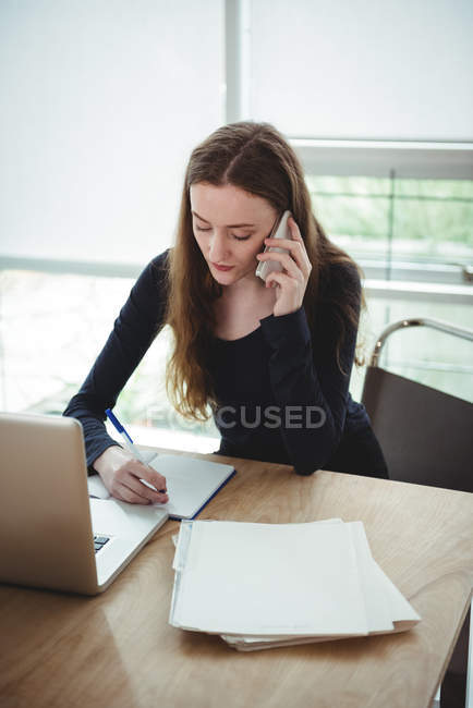 Business executive writing on diary while talking on mobile phone in office — Stock Photo