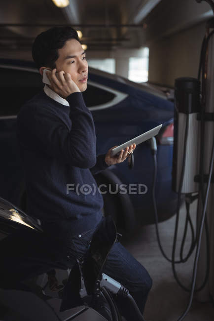 Мужчина разговаривает по мобильному телефону во время зарядки электромобиля в гараже — стоковое фото
