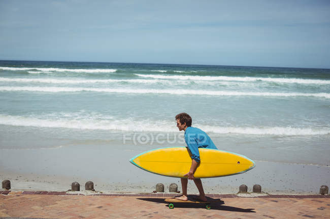 Hombre llevando tabla de surf mientras monta monopatín en la playa - foto de stock