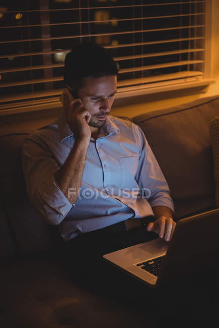 Чоловік розмовляє на мобільному телефоні, використовуючи ноутбук у вітальні вдома — стокове фото