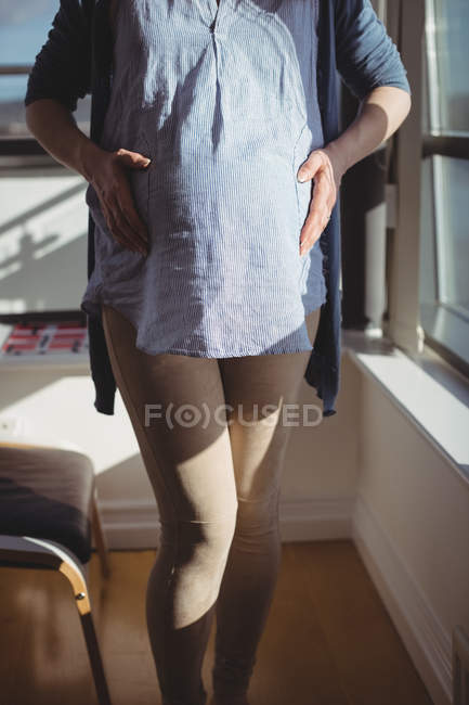 Средняя секция беременной женщины, стоящей у окна в гостиной дома — стоковое фото