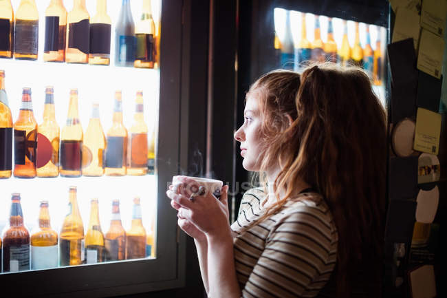 Mujer sosteniendo la taza de café y mirando la exhibición de vino en el bar - foto de stock