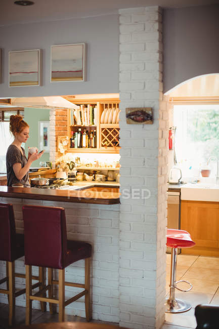 Жінка використовує мобільний телефон, маючи каву на кухні вдома — стокове фото