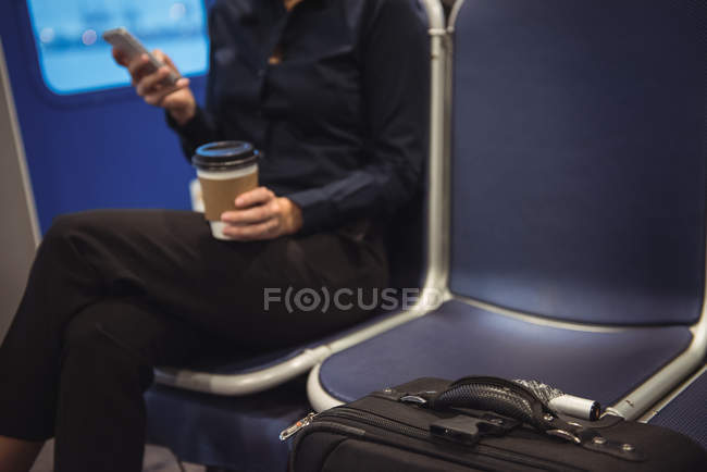 Sección media de la mujer de negocios con taza de café y equipaje usando el teléfono mientras está sentado en el tren - foto de stock