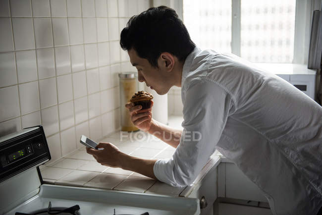 Чоловік використовує мобільний телефон, маючи кекс вдома — стокове фото