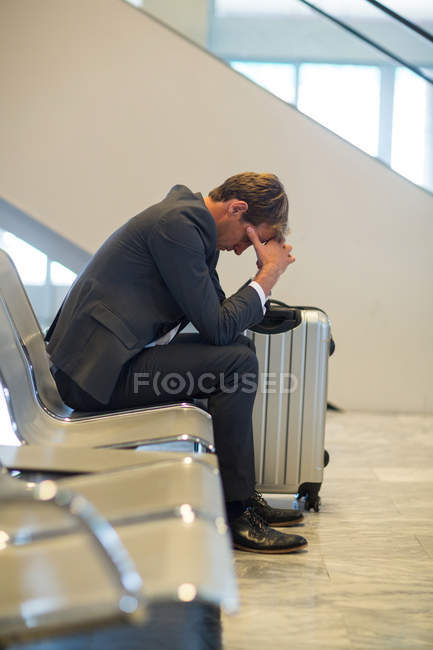 Tenso hombre de negocios sentado en la zona de espera con equipaje en la terminal del aeropuerto - foto de stock