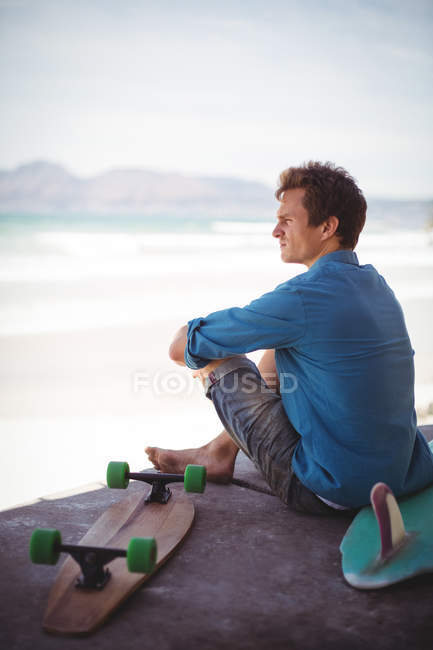 Hombre reflexivo con monopatín y tabla de surf sentado en la playa - foto de stock