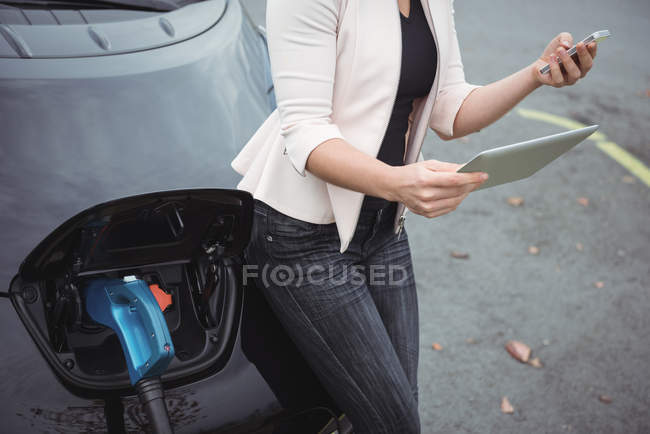 Partie médiane de la femme utilisant un téléphone portable tout en rechargeant la voiture électrique dans la rue — Photo de stock