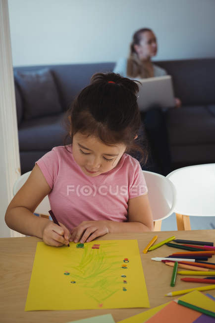 Menina atenta desenho em papel, enquanto a mãe usando laptop no fundo em casa — Fotografia de Stock