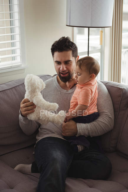 Father and baby playing with teddy bear on sofa in living room at home — Stock Photo