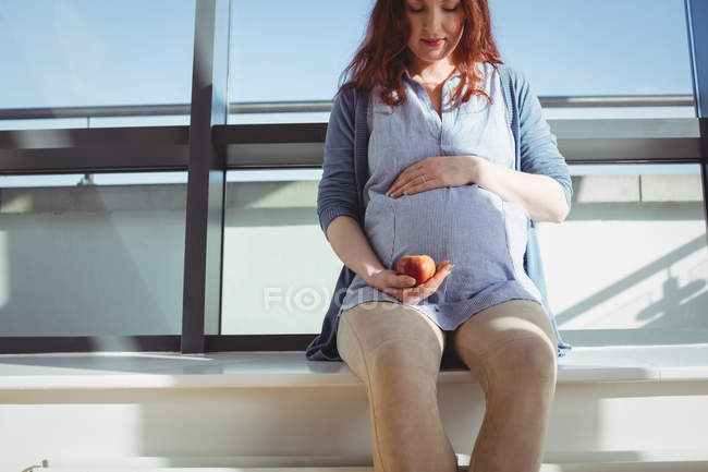 Mujer embarazada pensativa sentada cerca de la ventana y sosteniendo una manzana en casa - foto de stock