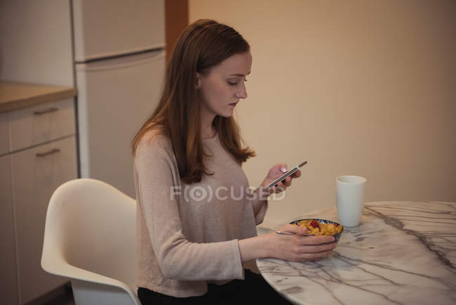 Жінка використовує мобільний телефон під час сніданку на кухні вдома — стокове фото