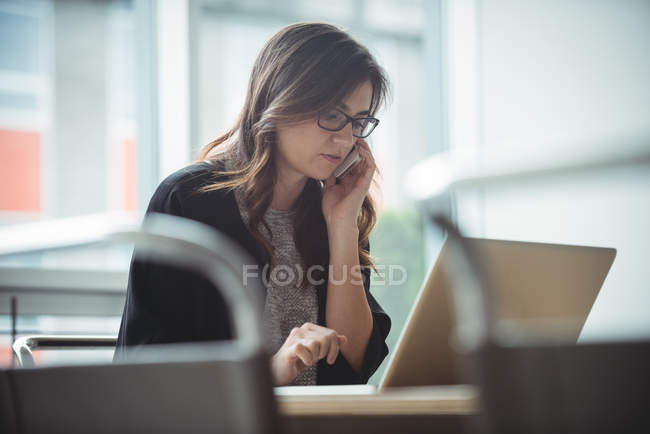 Ejecutivo de negocios hablando en el teléfono móvil mientras usa el ordenador portátil en la oficina - foto de stock