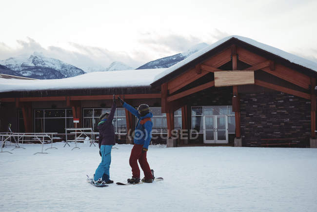 Couple snowboarding while high fiving on snow covered field by house — Stock Photo