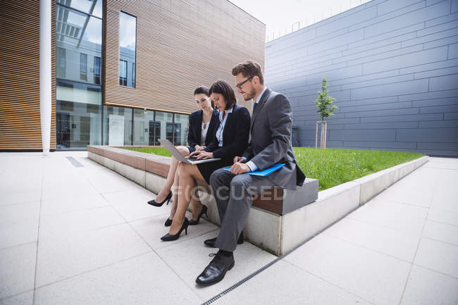 Empresaria sentada con colegas y usando laptop fuera del edificio de oficinas - foto de stock