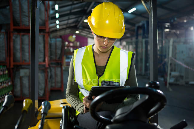 Travailleur féminin utilisant une tablette numérique assis sur un chariot élévateur dans un entrepôt — Photo de stock