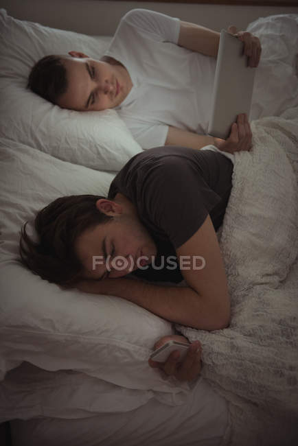 Гей пара використовує мобільний телефон і цифровий планшет, лежачи на ліжку в спальні — стокове фото