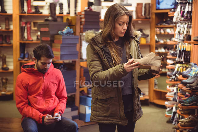 Mujer seleccionando zapato en una tienda mientras el hombre utiliza el teléfono móvil - foto de stock