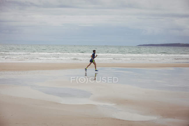 Atleta masculino correndo ao longo da praia molhada arenosa — Fotografia de Stock