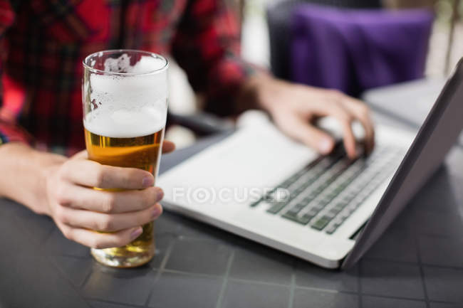 Seção média do homem usando laptop com copo de cerveja na mesa no bar — Fotografia de Stock