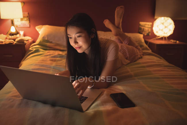 Женщина лежит и использует ноутбук на кровати в спальне — стоковое фото