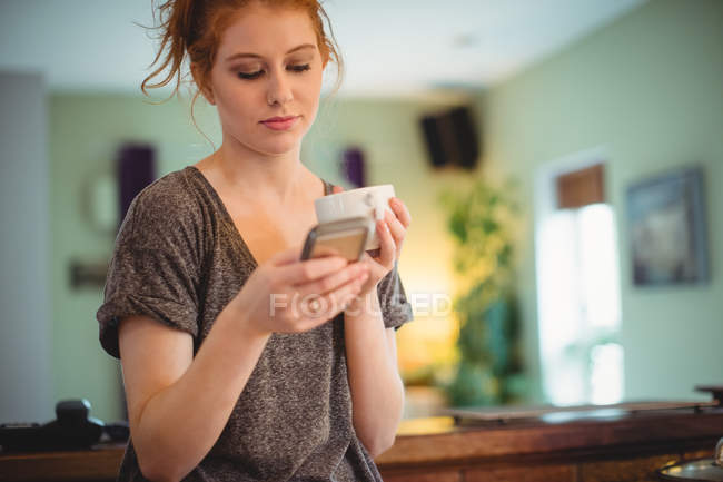 Beautiful woman using mobile phone while having coffee in kitchen at home — Stock Photo