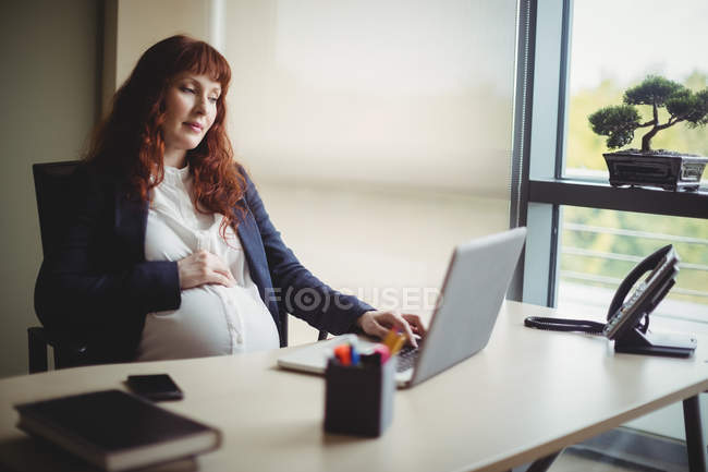 Вагітна бізнес-леді тримає живіт при використанні ноутбука в офісі — стокове фото