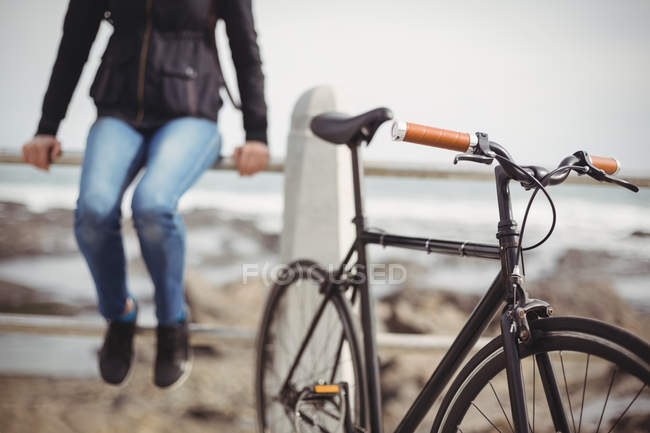 Vélo debout près du bord de mer avec une femme assise sur une rampe — Photo de stock