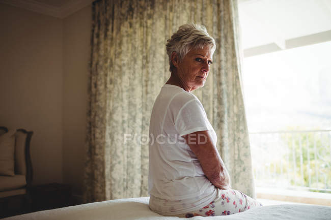 Thoughtful senior woman sitting on a bed in bedroom at home — Stock Photo