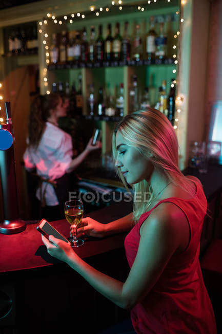 Mujer usando el teléfono móvil mientras toma una copa de vino en el mostrador en el bar - foto de stock