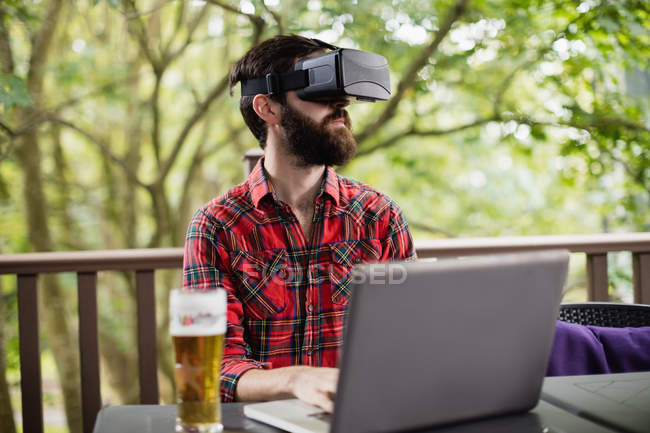 Homem usando fone de ouvido de realidade virtual ao usar laptop no bar — Fotografia de Stock