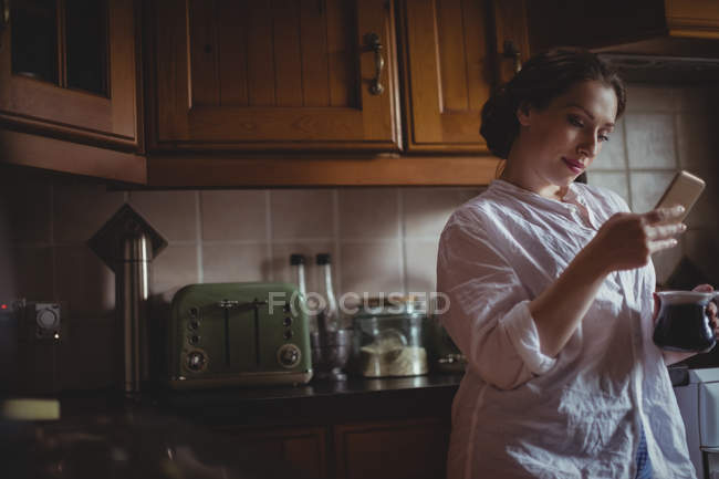 Belle femme utilisant un téléphone portable tout en prenant un café dans la cuisine à la maison — Photo de stock