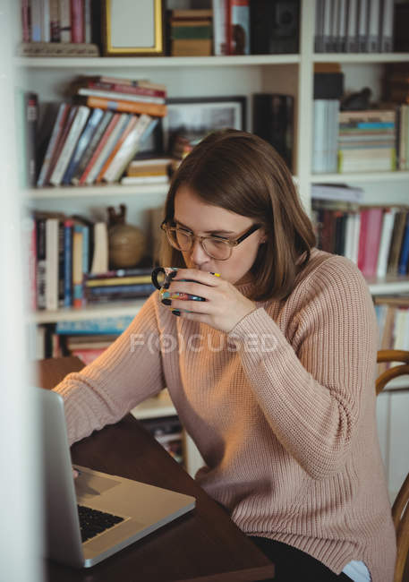 Mujer usando el ordenador portátil mientras bebe café en la sala de estar en casa - foto de stock