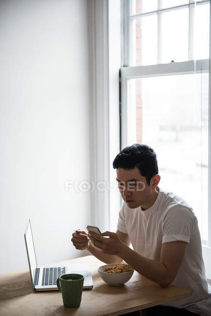 Homem usando telefone celular enquanto toma café da manhã em casa — Fotografia de Stock