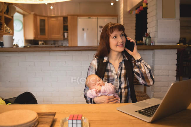 Mother carrying baby while talking on mobile phone at home — Stock Photo