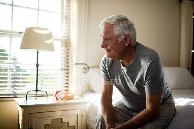 Задумчивый старший мужчина сидит на кровати в спальне дома — стоковое фото