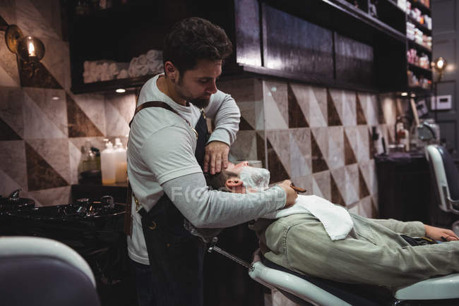 Homme se faire raser la barbe par un coiffeur avec rasoir dans un salon de coiffure — Photo de stock