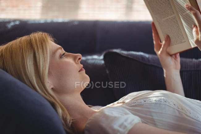 Beautiful woman lying on sofa and reading book in living room at home — Stock Photo